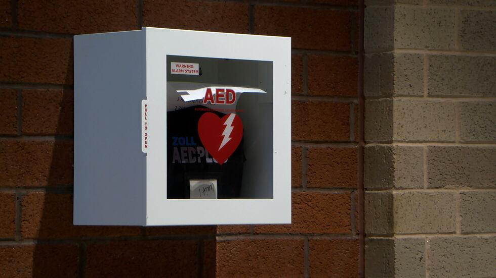 An Automated External Defibrillator (AED) is mounted on the wall in a school. The device plays...